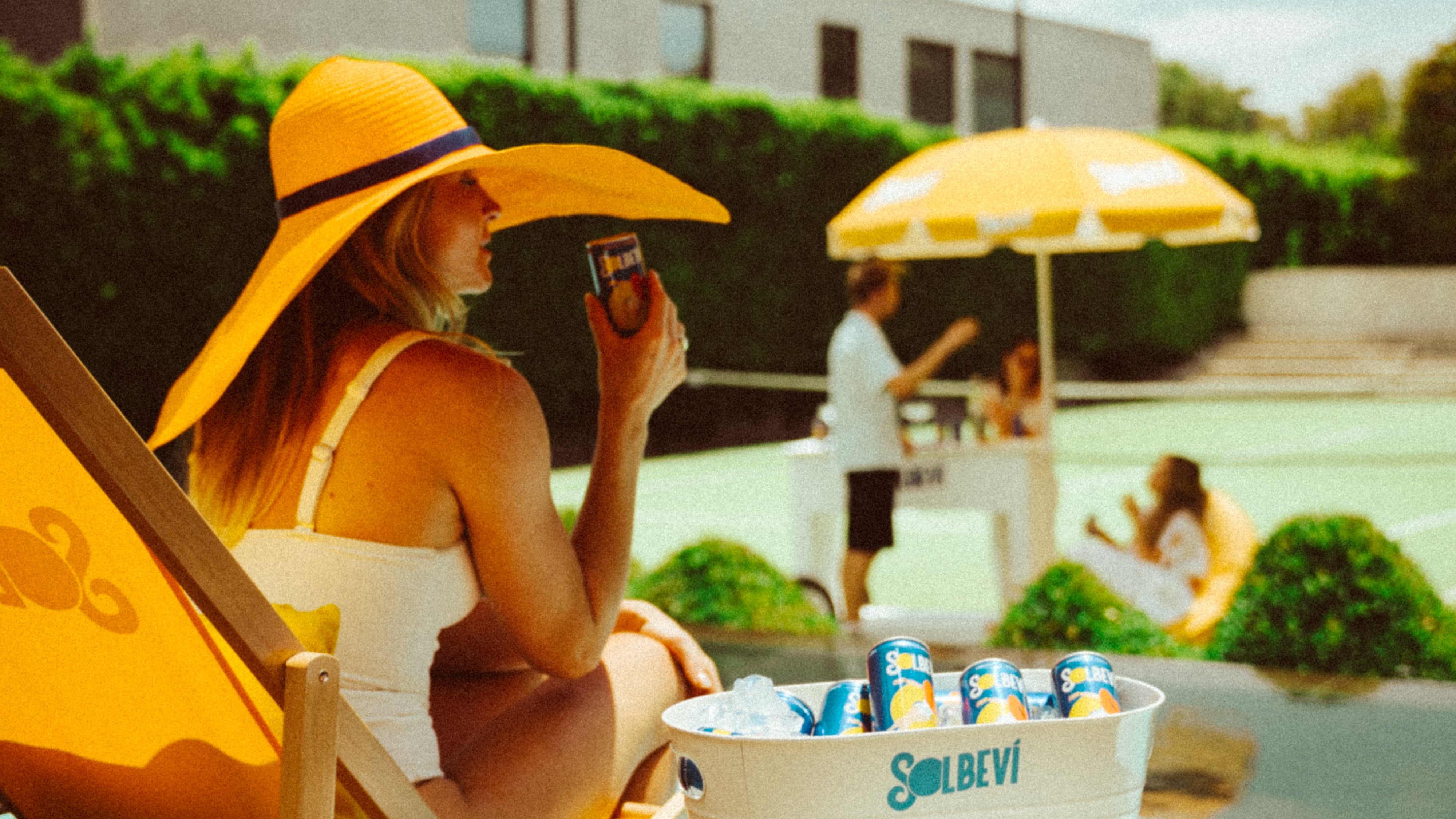 Woman in a yellow hat and dress sitting by a pool with a cooler of drinks on a table.